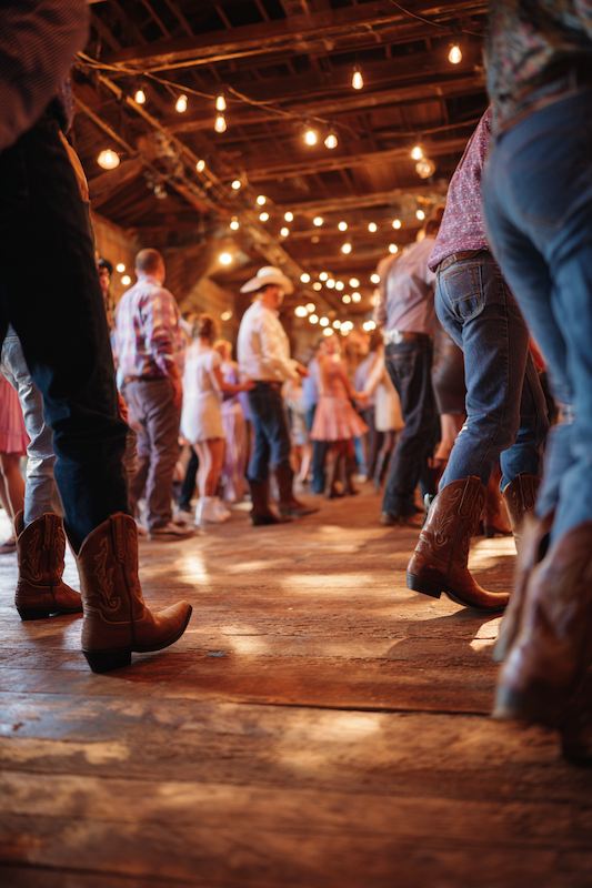Guests in cowboy boots dancing at a rustic cowboy wedding reception, illuminated by warm string lights in a barn-like venue