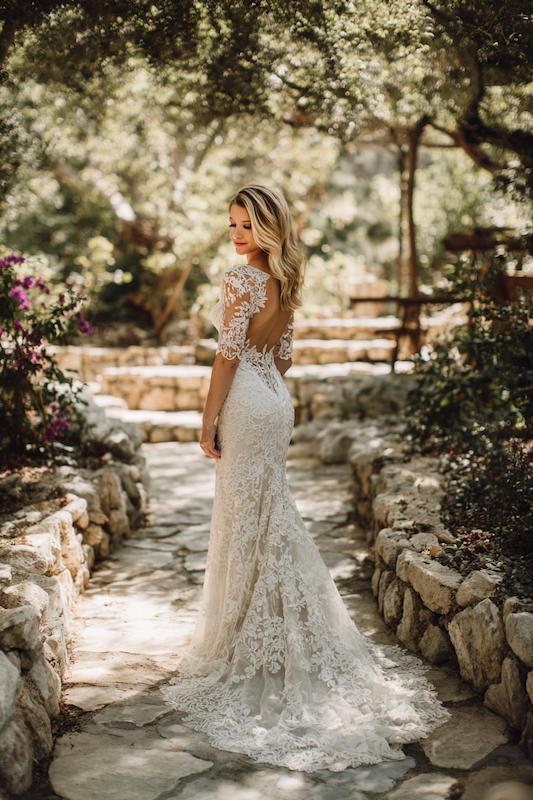 Lace mermaid wedding dress A bride in a lace mermaid wedding dress with sheer, long sleeves, and a low back, standing in a garden setting with stone pathways and greenery