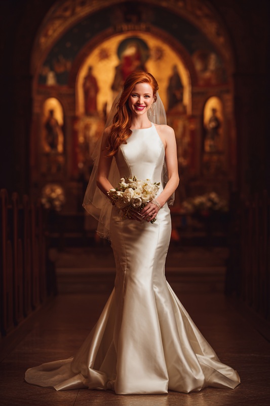 Satin – The Classic Choice for mermaid wedding dresses A bride in a sleek satin mermaid wedding dress with a high neckline, holding a bouquet of white flowers, and walking down the aisle in a beautiful church
