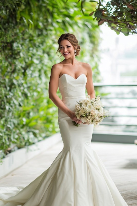 today's mermaid wedding dress A bride smiling in a sleek, strapless mermaid wedding dress, holding a bouquet of white flowers, with a lush green backdrop