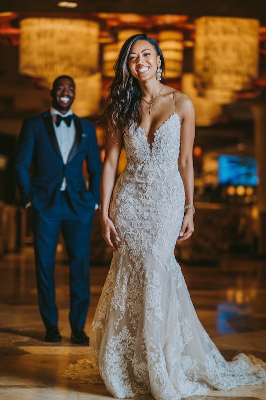 bride in a mermaid wedding dress with groom in the background Bride in a fitted mermaid wedding dress with intricate lace detailing, smiling joyfully with her groom in a tuxedo walking behind her, set against a glamorous background