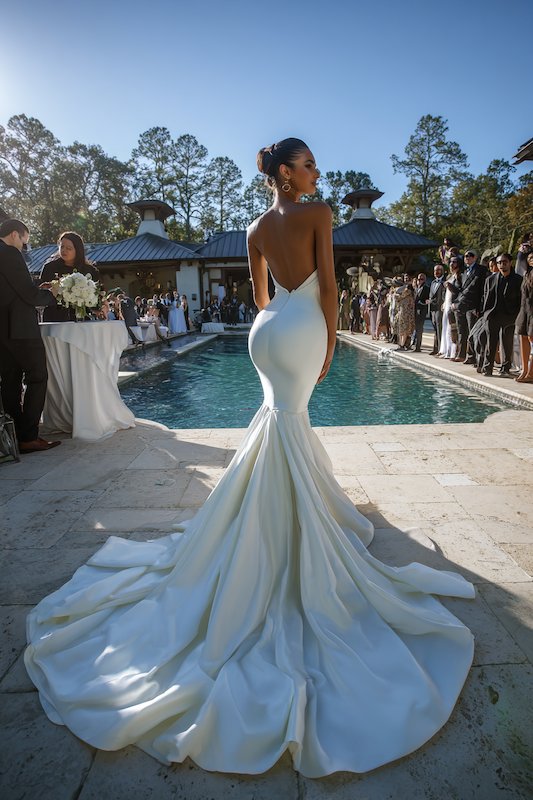 pros of a mermaid wedding dress A bride in a white satin mermaid wedding dress with an open back and a dramatic train, standing by a poolside at a wedding reception
