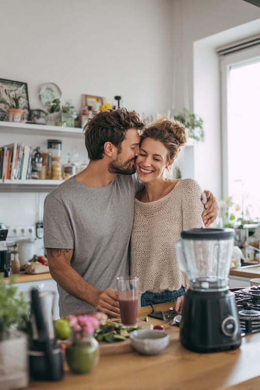 Couple sharing a sweet kiss in a cozy kitchen while making smoothies together, with a black blender and fresh ingredients on the counter.