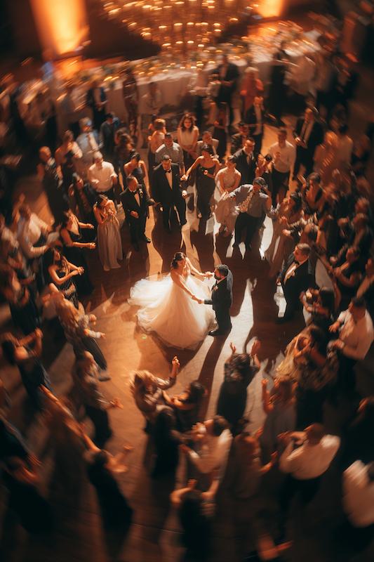 Bride and groom share a romantic first dance surrounded by guests in a glowing circle under warm chandelier light.
