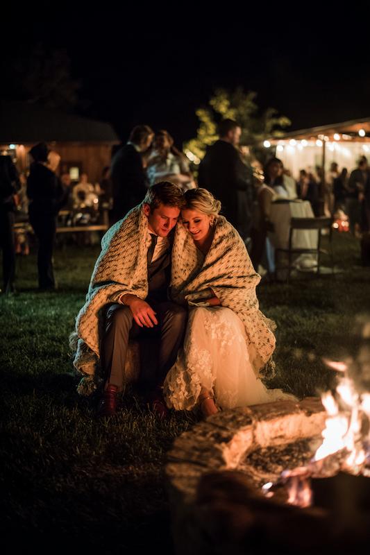 Bride and groom wrapped in a blanket share a cozy moment by the fire pit during their outdoor night wedding reception.