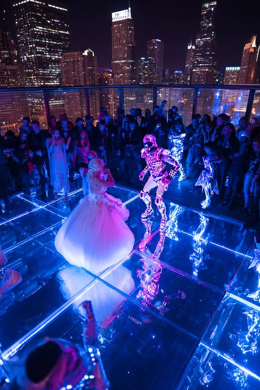 Bride dancing with LED robot performer on a glowing rooftop dance floor overlooking a city skyline at night.