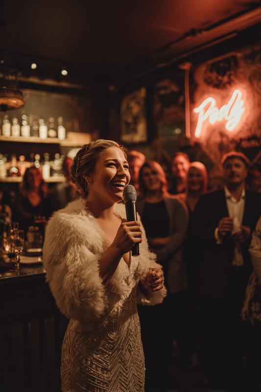 Bride in a beaded gown with a fur wrap giving a joyful toast at a warmly lit wedding reception bar.