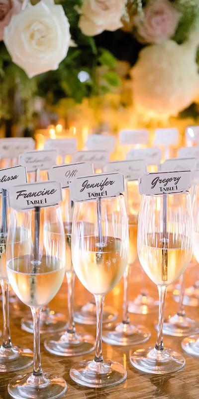 Champagne glasses with escort cards displaying guest names and table numbers, set against candles and flowers.