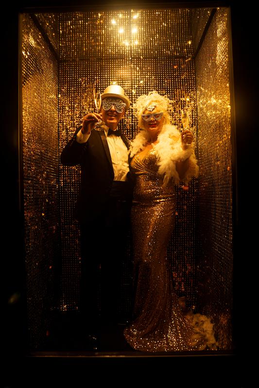 Couple in glamorous attire posing in a gold sequin photo booth, wearing playful masks and holding champagne glasses, creating a festive and celebratory vibe