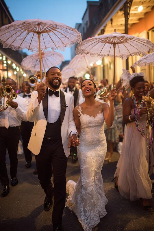 Newlywed couple leads a joyful second line parade, holding lace parasols as a brass band and guests celebrate in the street.