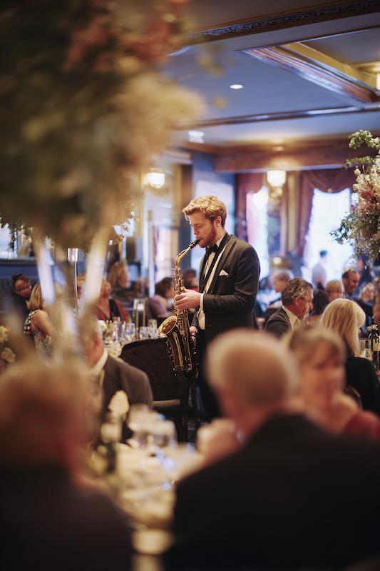 Saxophonist in a tuxedo performing live during a black-tie wedding reception, surrounded by elegantly dressed guests seated at candlelit dinner tables