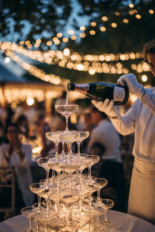 Waiter in white gloves pouring champagne into a tall glass pyramid during a wedding toast.