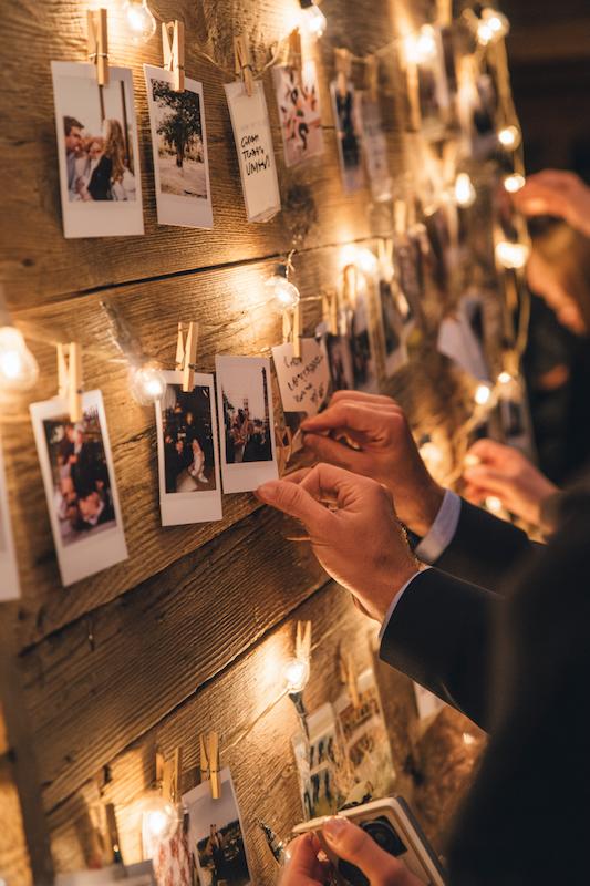 Wedding guest pinning Polaroid photos to a wooden board with string lights as a creative guestbook alternative.