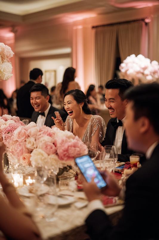 Wedding guests laughing and enjoying dinner at a reception table decorated with pink peony centerpieces and glowing candles.