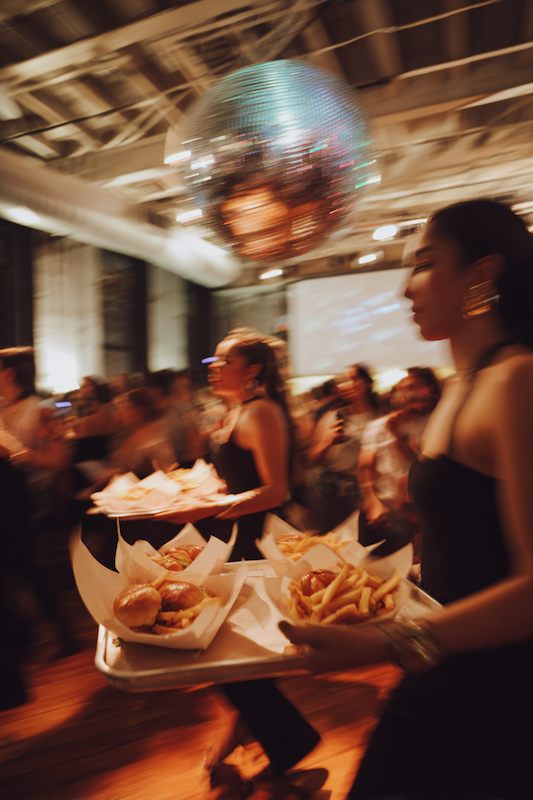 Wedding servers carrying trays of late-night sliders and fries across a lively dance floor under a disco ball.