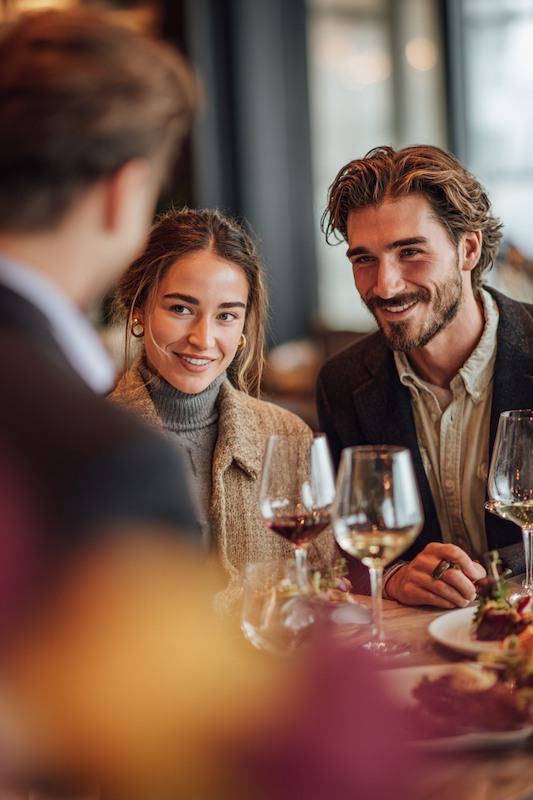 Wedding Tasting: What You Need to Know 1 Couple smiling during a wedding tasting, seated at a table with wine glasses and plated dishes, speaking with a server