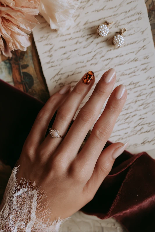 A close-up of a hand resting on handwritten paper, showcasing a pearl ring and a tortoiseshell-patterned accent nail, with lace sleeve detail and pearl earrings nearby A close-up of a hand resting on handwritten paper, showcasing a pearl ring and a tortoiseshell-patterned accent nail, with lace sleeve detail and pearl earrings nearby