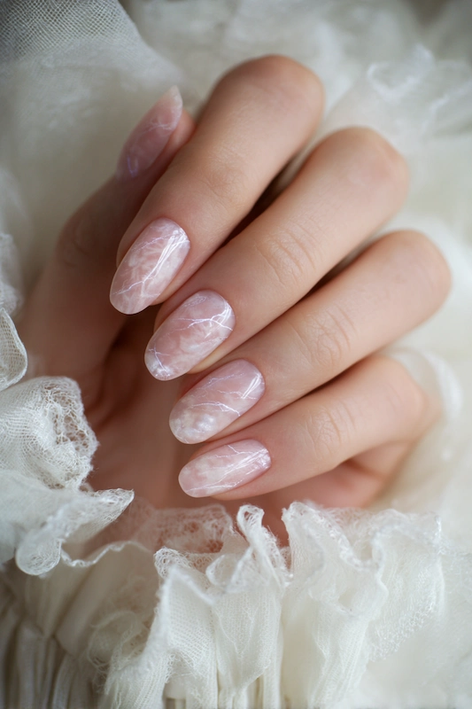 Close-up of a hand with long, almond-shaped nails featuring a soft pink marble design, resting against delicate white lace fabric Close-up of a hand with long, almond-shaped nails featuring a soft pink marble design, resting against delicate white lace fabric