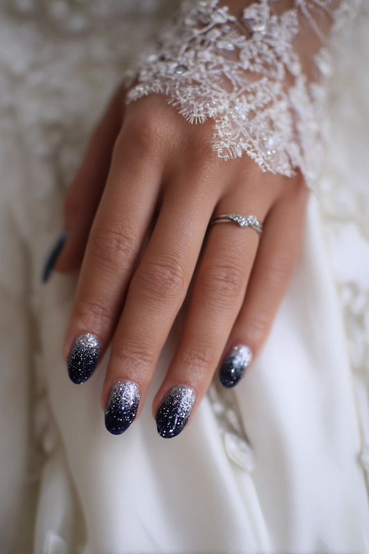 Close-up of a hand with navy-to-silver glitter ombré nails, adorned with a silver ring and resting on white fabric with lace sleeve detailing Close-up of a hand with black-to-silver glitter ombré nails, adorned with a silver ring and resting on white fabric with lace sleeve detailing