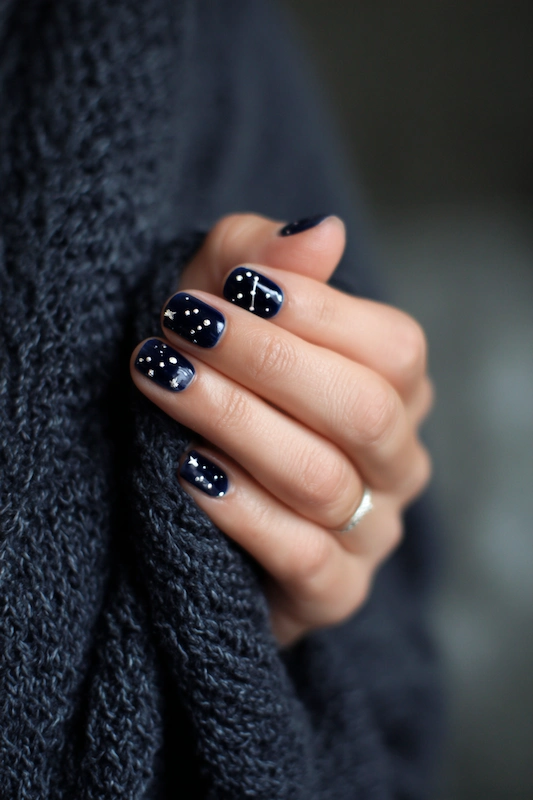 Close-up of a hand with short navy blue nails decorated with white star constellations, resting against a dark knit fabric Close-up of a hand with short navy blue nails decorated with white star constellations, resting against a dark knit fabric