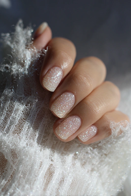 Close-up of a hand with short, square-shaped nails painted in a sheer nude shade with fine silver glitter, resting against a shimmery white knit fabric Close-up of a hand with short, square-shaped nails painted in a sheer nude shade with fine silver glitter, resting against a shimmery white knit fabric