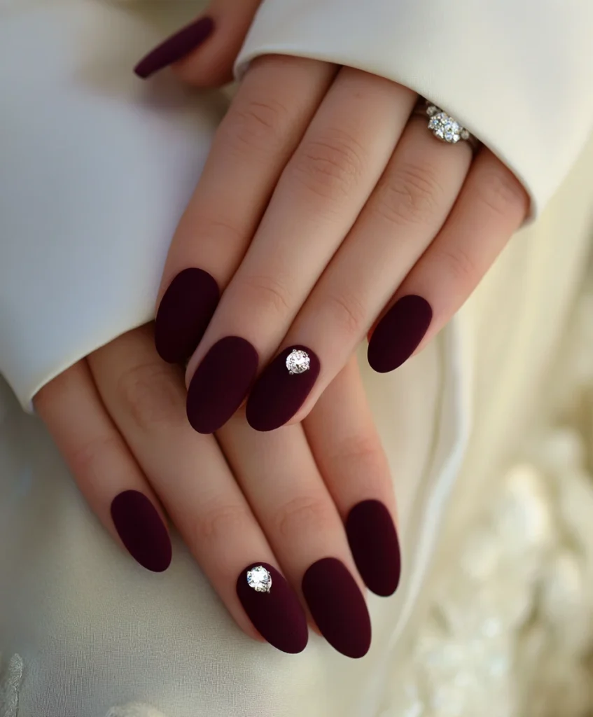 Close-up of hands with matte burgundy almond-shaped nails, each featuring a single rhinestone accent, paired with a sparkling diamond ring and white satin sleeves. Close-up of hands with matte burgundy almond-shaped nails, each featuring a single rhinestone accent, paired with a sparkling diamond ring and white satin sleeves.