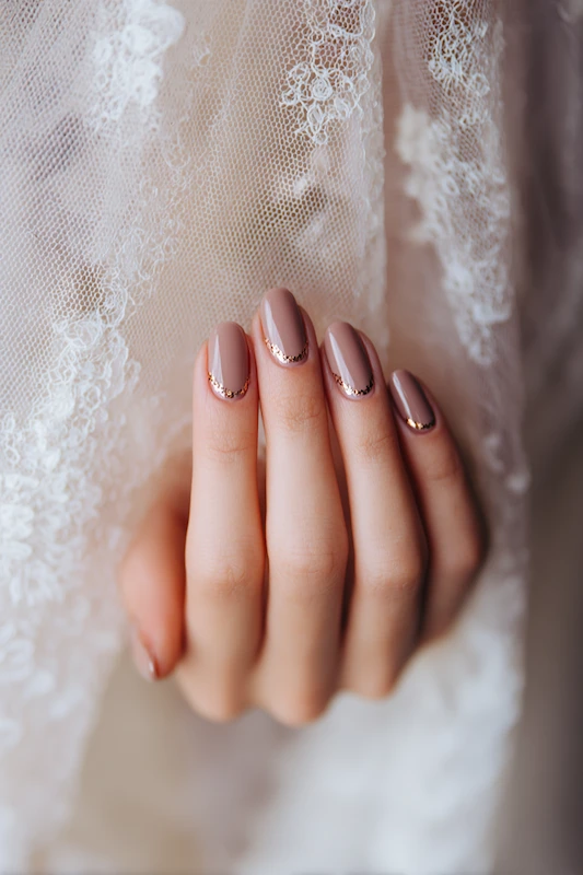 close-up of a hand with mauve-pink polished nails featuring delicate gold glitter accents near the cuticles, resting gently against sheer white lace fabric with floral embroidery. close-up of a hand with mauve-pink polished nails featuring delicate gold glitter accents near the cuticles, resting gently against sheer white lace fabric with floral embroidery.