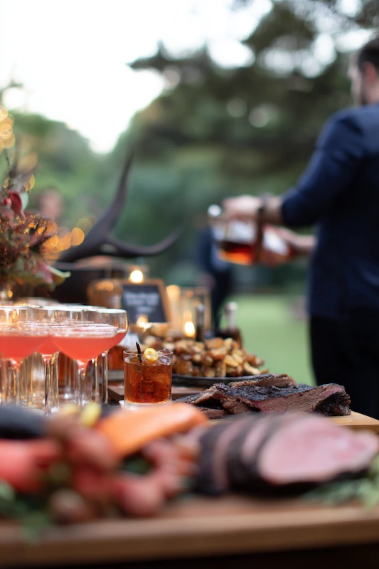 25 Wedding BBQ Buffet Ideas That Instantly Upgrade Your Reception 18 BBQ buffet with cocktail pairings featuring sliced brisket, roasted veggies, and bourbon pours at an elegant garden reception