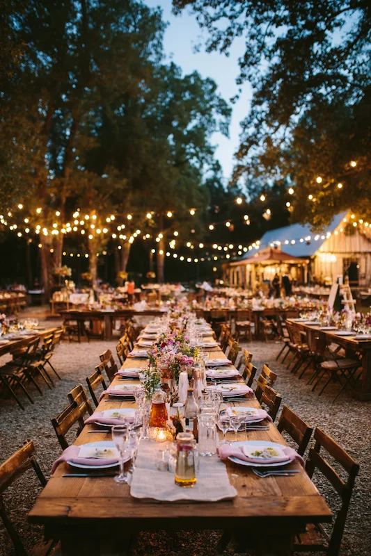 25 Wedding BBQ Buffet Ideas That Instantly Upgrade Your Reception 5 A beautifully lit outdoor BBQ wedding setup with long wooden tables, flowers, glassware, and string lights creating a warm, inviting atmosphere