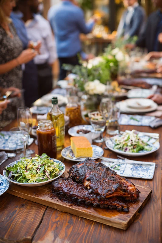 25 Wedding BBQ Buffet Ideas That Instantly Upgrade Your Reception 4 Wedding BBQ Buffet featuring tender ribs, salad, cornbread, and sauces on a wooden table surrounded by elegant glassware and guests