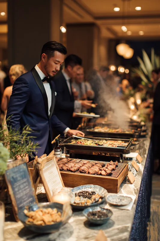 25 Wedding BBQ Buffet Ideas That Instantly Upgrade Your Reception 23 Groom in midnight blue tux selects from BBQ buffet with steam rising, chalkboard signs, and guests mingling in a ballroom