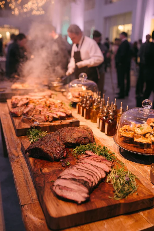 25 Wedding BBQ Buffet Ideas That Instantly Upgrade Your Reception 25 Indoor wedding smokehouse station with steaming carved meats, chef in background, mini sauce bottles, and cloche-covered sides on wooden boards