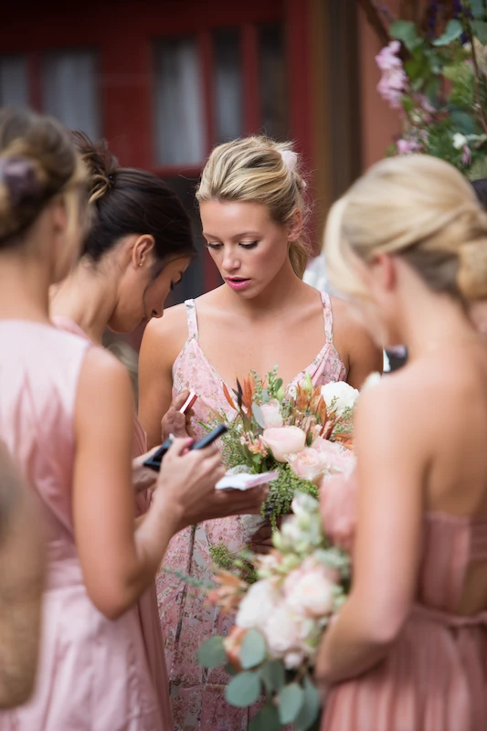 Maid Of Honor Duties: Your No Stress Game Plan 1 Bridesmaids in soft pink dresses gather closely as one woman checks a small notebook while holding a bouquet of blush and greenery flowers during a wedding prep moment.