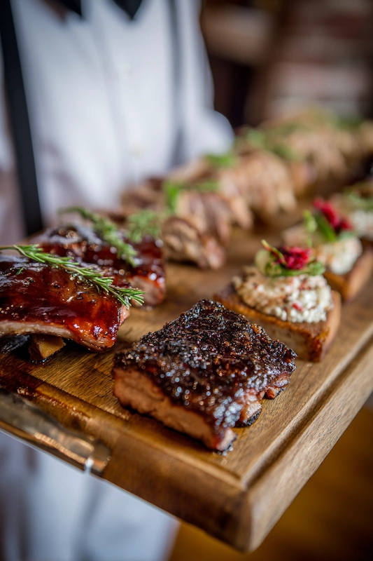 25 Wedding BBQ Buffet Ideas That Instantly Upgrade Your Reception 8 A wooden tray with BBQ ribs, grilled meats, and garnishes, served by a waiter in a tuxedo, offering a regional BBQ tasting