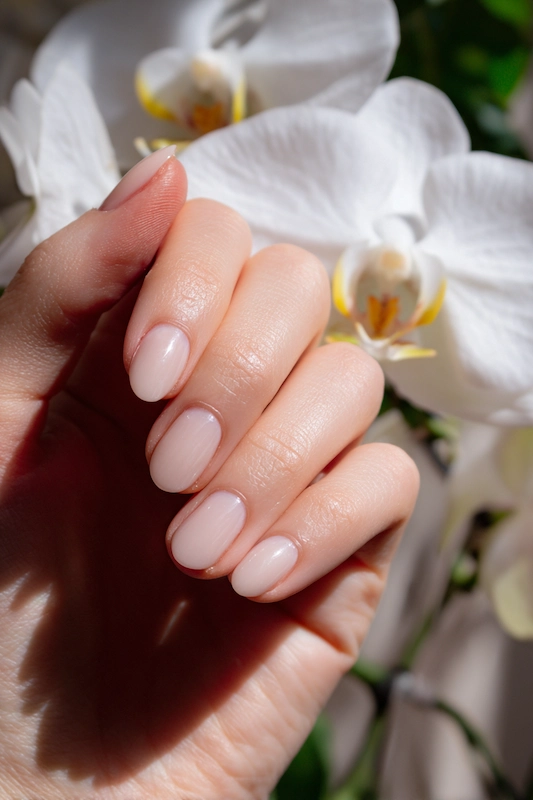 Beyond the Almond: The 2026 Engagement Nail Trends Defining "Intentional Glamour" 11 Short, Natural, Old Money Nails in soft nude on a hand posed before white orchids, glossy, clean engagement-ready look.