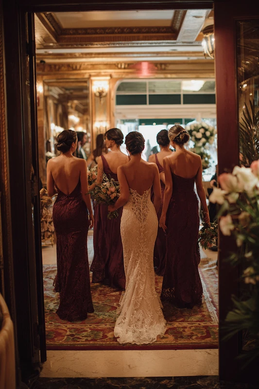 Maid Of Honor Duties: Your No Stress Game Plan 8 Bride in a lace gown walks with her bridesmaids in deep burgundy dresses through an ornate hallway toward the ceremony, captured from behind in warm, elegant lighting.
