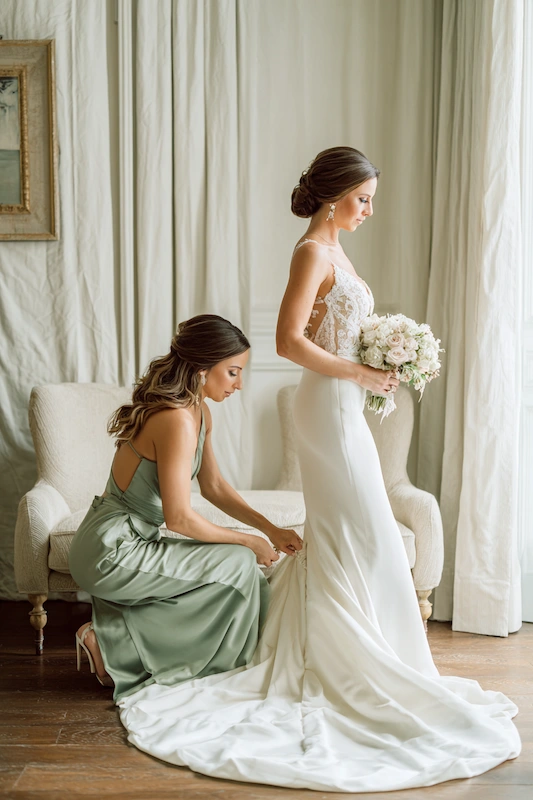 Maid Of Honor Duties: Your No Stress Game Plan 5 Bride standing in a fitted white gown holding a soft blush bouquet while a bridesmaid in a sage dress kneels behind her, adjusting the train in a bright, elegant room.