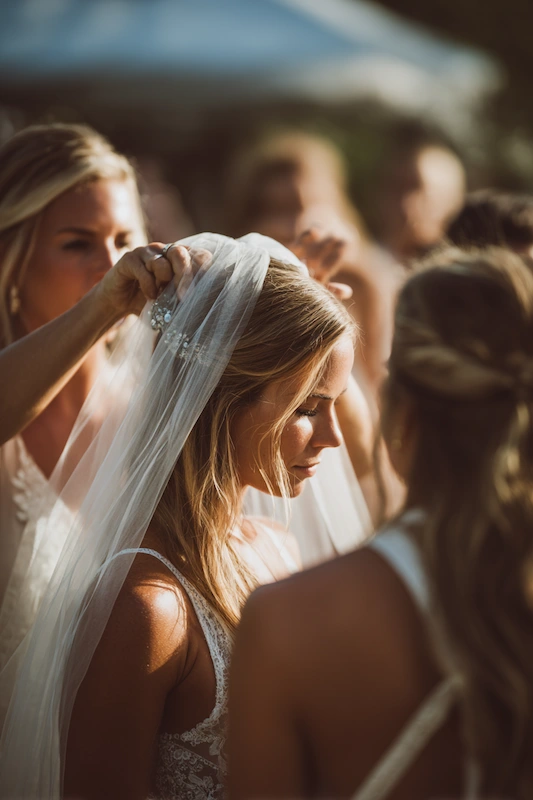 Maid Of Honor Duties: Your No Stress Game Plan 10 Bride standing in soft golden light as a bridesmaid gently adjusts her veil, surrounded by women in a serene outdoor wedding moment.