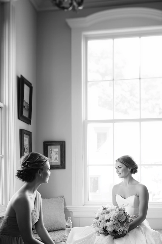Maid Of Honor Duties: Your No Stress Game Plan 2 Bride sitting by a large bright window holding a bouquet of light roses, warmly talking with a bridesmaid seated nearby in a serene black-and-white getting-ready room.