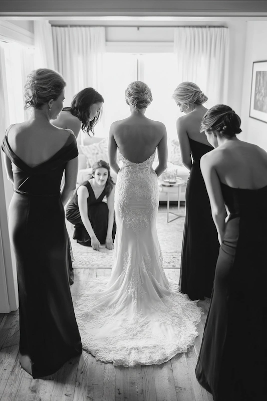 Maid Of Honor Duties: Your No Stress Game Plan 14 Black and white scene of a bride in a fitted lace gown standing in the center while her bridesmaids in dark dresses gather around to adjust her train and help her get ready