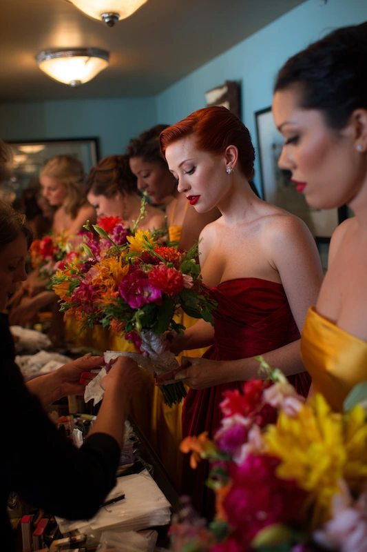 Maid Of Honor Duties: Your No Stress Game Plan 9 Bridesmaids in bold red and yellow dresses hold vibrant mixed-color bouquets while preparing at a crowded makeup table, captured in warm indoor lighting.