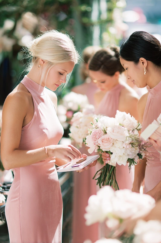 Maid Of Honor Duties: Your No Stress Game Plan 4 Bridesmaids in blush pink dresses gather together, one adjusting items on a small clipboard while another holds a bouquet of pale pink and white flowers in soft outdoor light.