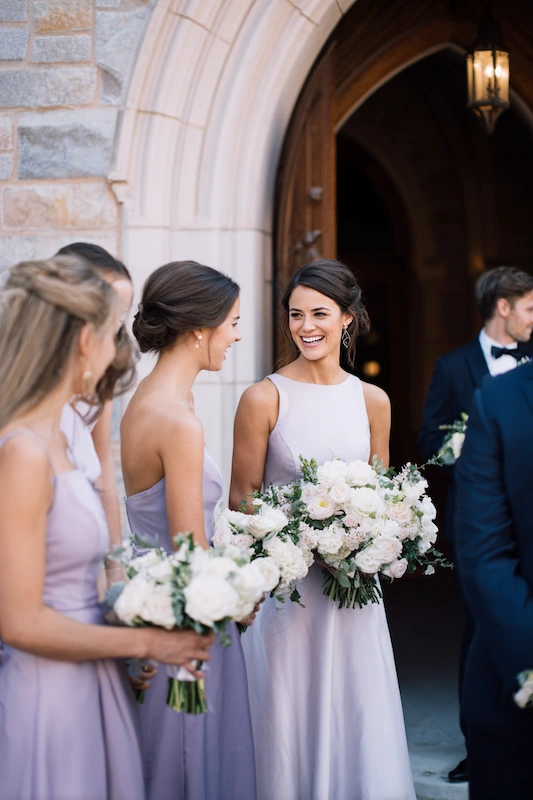 Maid Of Honor Duties: Your No Stress Game Plan 11 Bridesmaids in soft lavender dresses smiling and chatting outside a stone church entrance while holding elegant white and blush bouquets