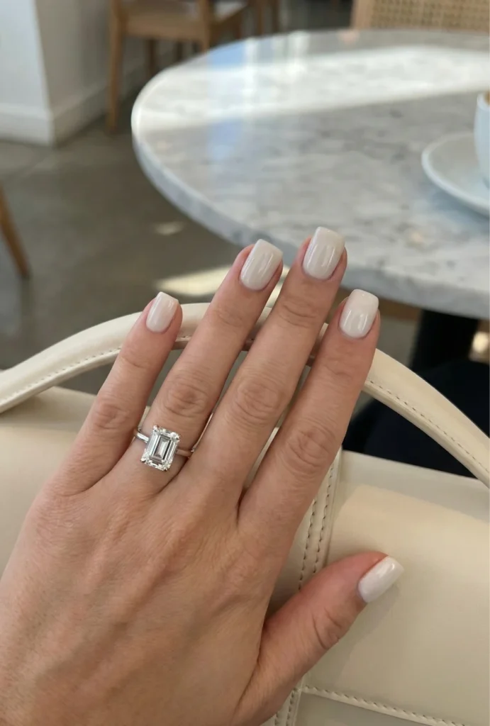Beyond the Almond: The 2026 Engagement Nail Trends Defining "Intentional Glamour" 6 Neutral nude square nails with emerald cut engagement ring resting on cream handbag at café table