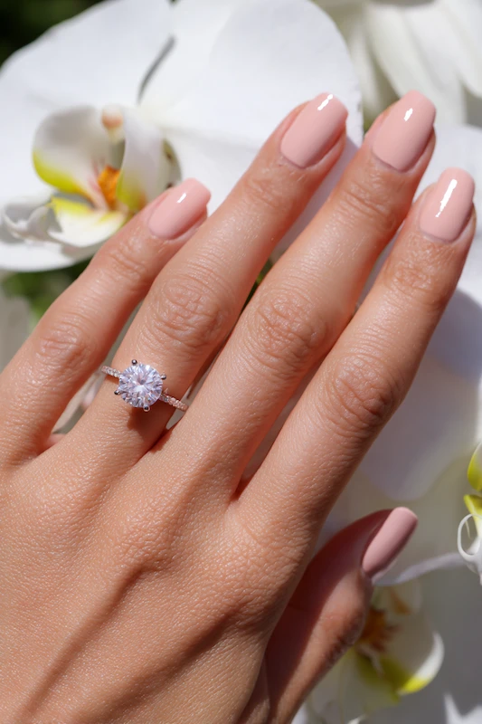 Beyond the Almond: The 2026 Engagement Nail Trends Defining "Intentional Glamour" 14 Peachy nude manicure with round diamond engagement ring against white orchids
