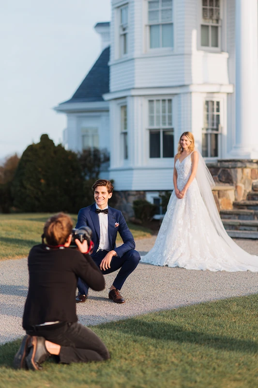 Old Money Wedding Aesthetic: Your Complete Style Guide 21 Photographer capturing groom kneeling as bride stands behind him outside a white coastal estate during golden hour