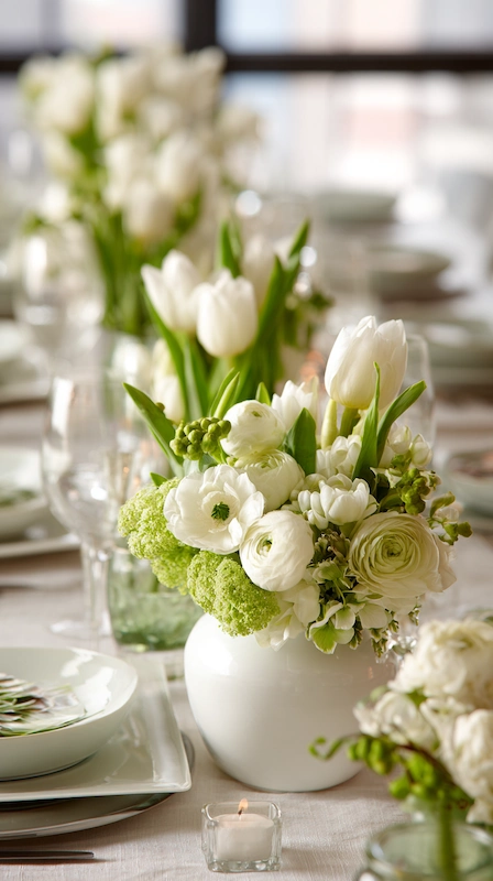 35 Spring Wedding Centerpieces With Flower Pairings Brides Can Actually Use 4 All white wedding table with tulips and ranunculus in bud vases, soft greenery accents, candlelight, and clean modern place settings
