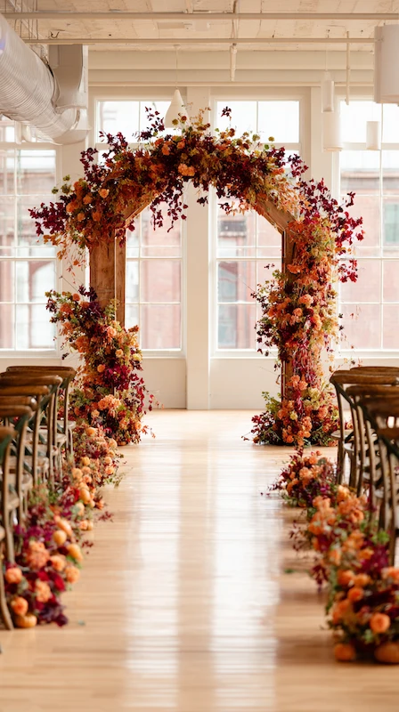 Autumn wedding ceremony aisle with warm fall florals, wooden arch, and rustic chairs in a bright loft with large windows.
