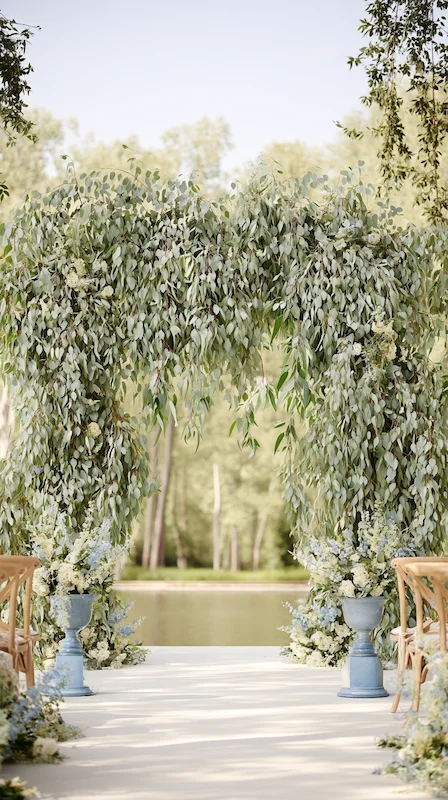 Eucalyptus wedding arch with cascading greenery and soft florals set beside a serene lakeside outdoor ceremony backdrop