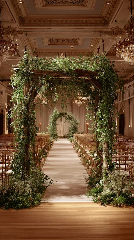 Elegant indoor wedding ceremony aisle with greenery-covered wooden arch, garden florals, and rows of chairs in a grand ballroom.